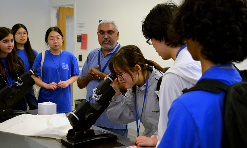 Students gathers around a lab bench to participate in a science activity while a student leans forward, looking intently through the eyepiece of a black laboratory microscope